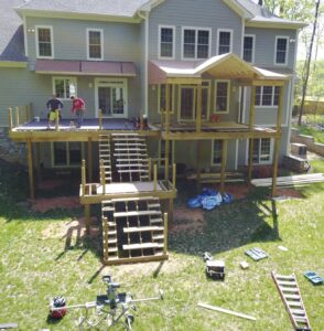 Two contractors are assembling a large wooden deck on the backside of a house on a sunny day.