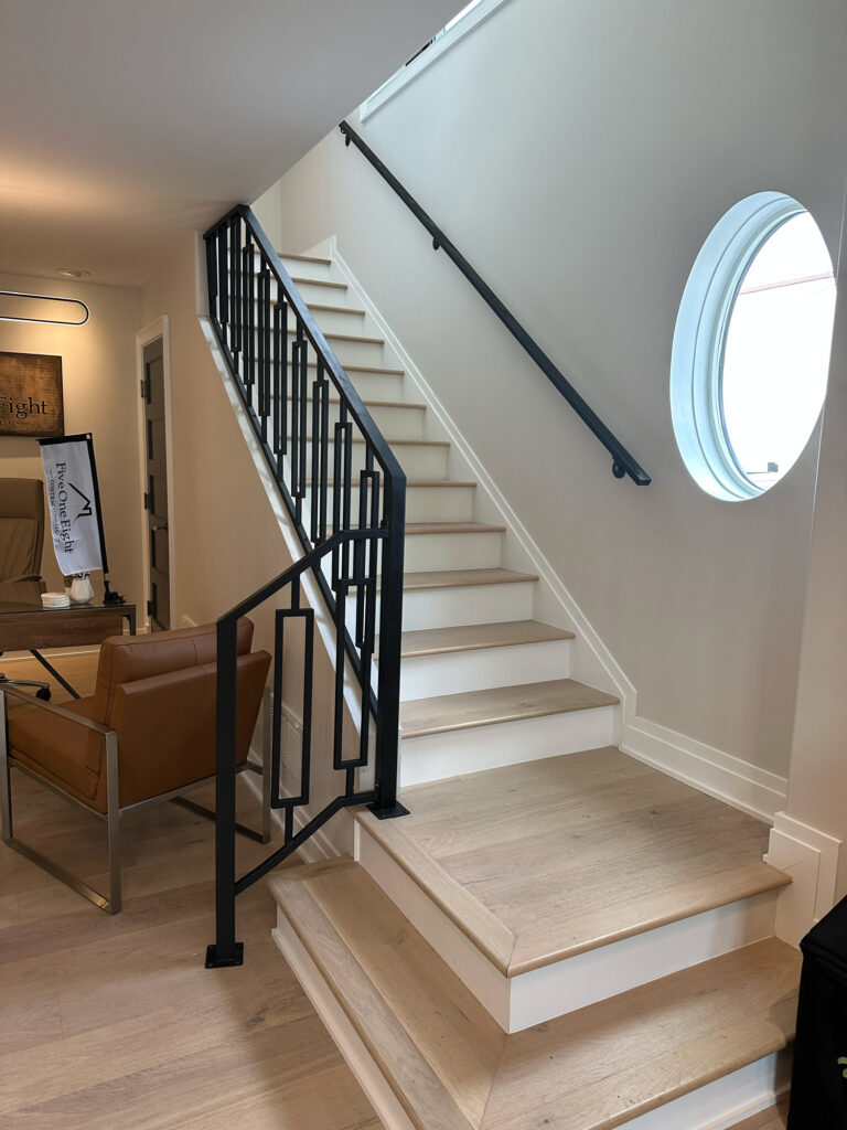 Looking up a modern set of stairs featuring geometrically pattern metal balusters, oval shaped window, and light beige flooring.