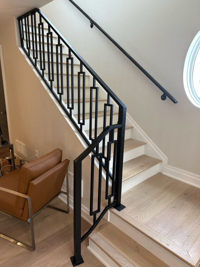 Looking up a modern set of stairs featuring geometrically pattern metal balusters, oval shaped window, and light beige flooring.