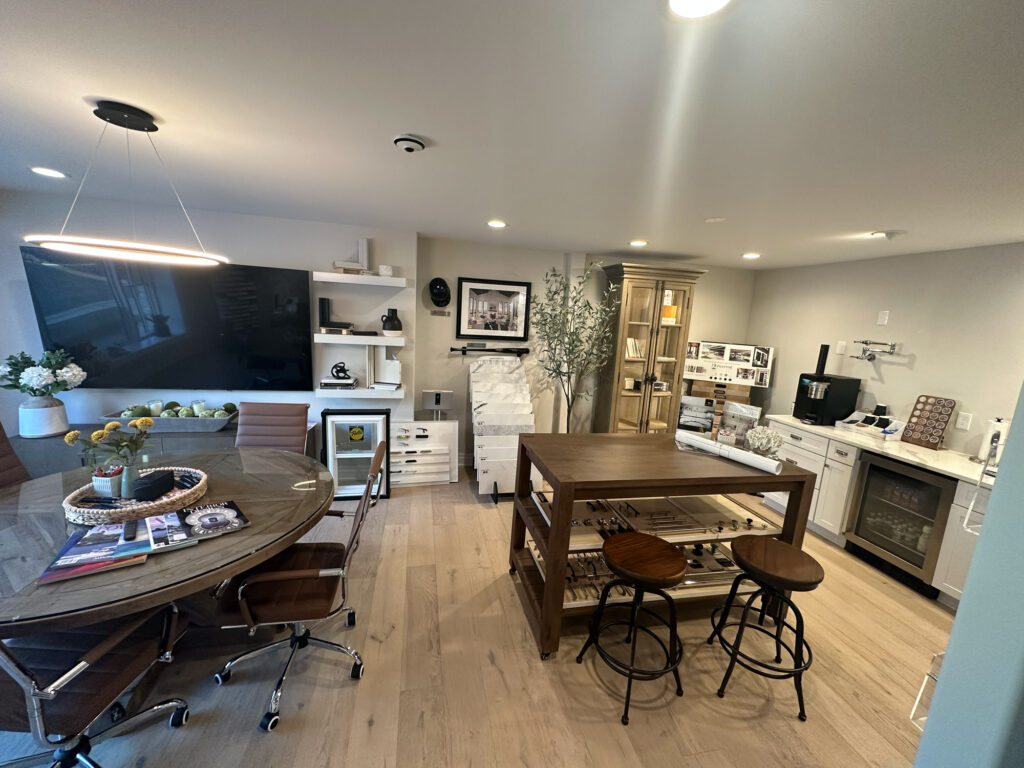 Modern office with sleek design featuring a large wooden table with chairs, wall shelves with decor, a TV, and kitchen area with light wood flooring.